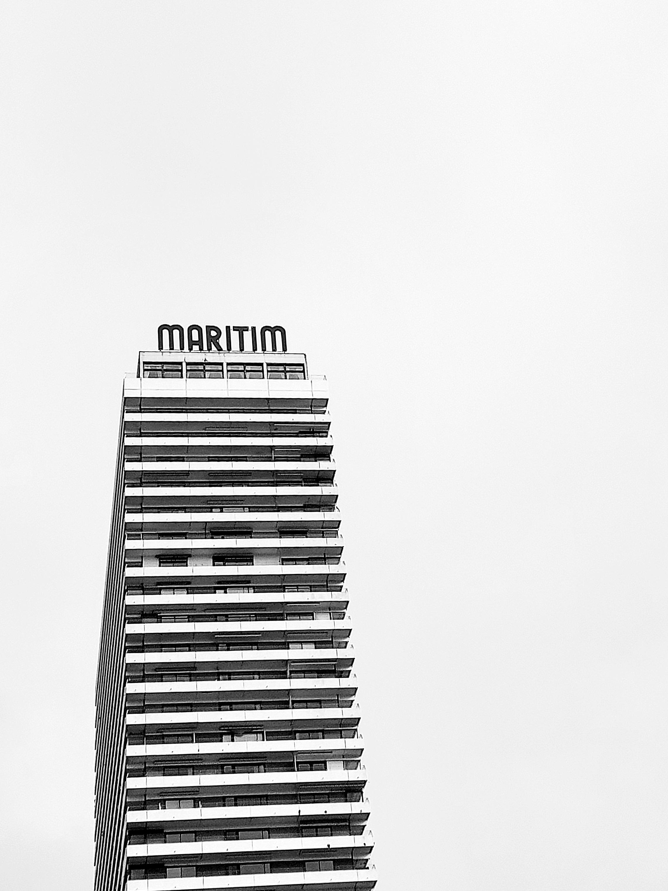 Black-and-white shot. A tall, slender building rises into the bright sky. Windows and wide balconies create a horizontal rhythm of light and dark. A double row of windows can be seen above the top floor. The building is crowned by huge, freestanding letters spelling out ‘MARITIM’.
A minimalist image for maximum holiday fun. Perhaps.