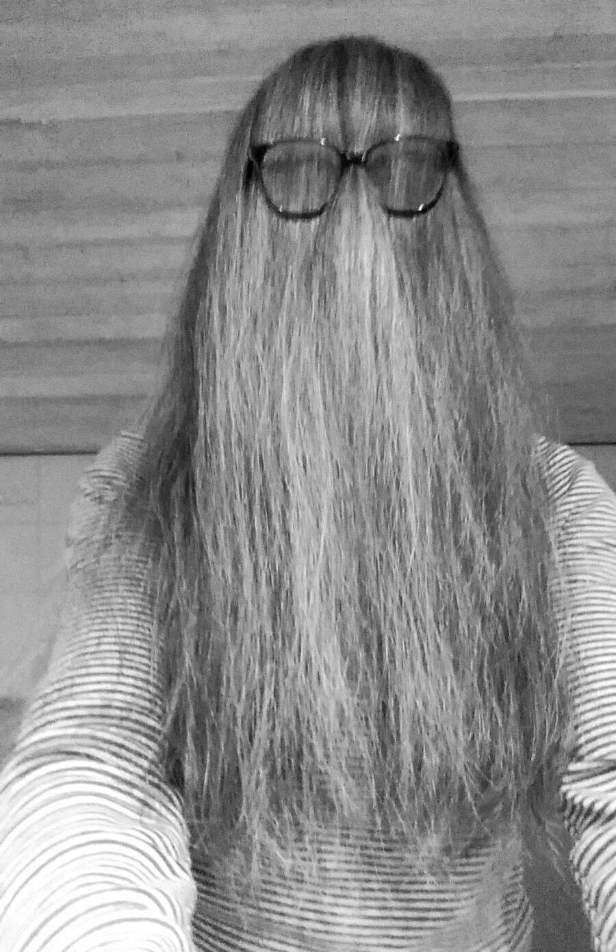 Black-and-white selfie. A person wearing a light-and-dark-striped longsleeve has combed their long hair forwards, completely covering their face and chest. A pair of glasses with clear lenses sits on their hair at eye level.