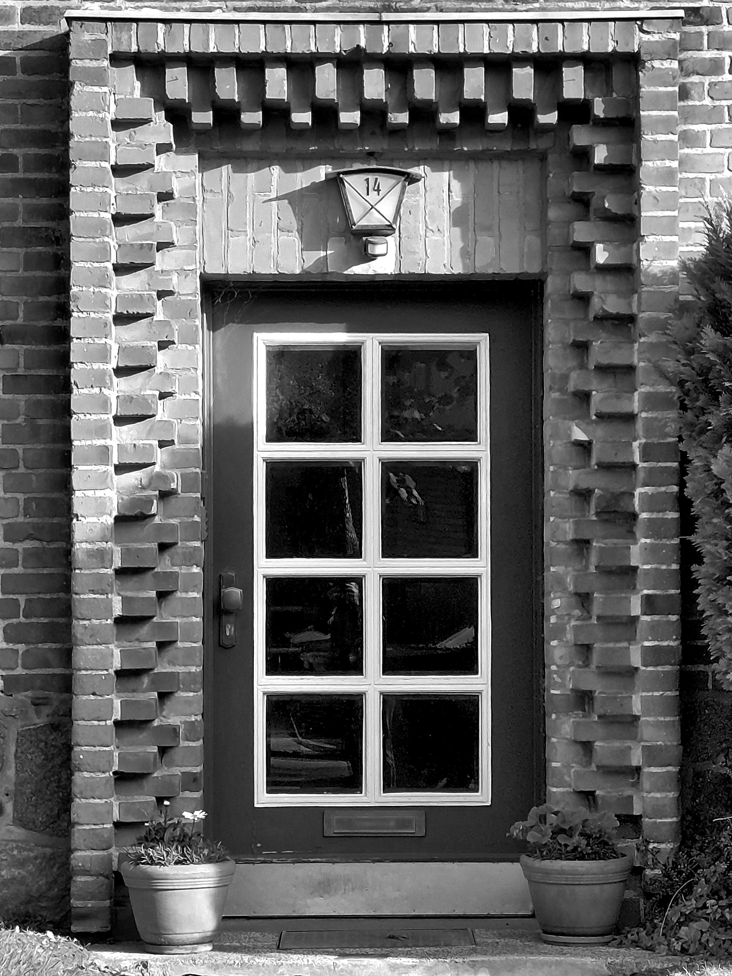 Black-and-white capture. A panelled door with eight glass panes, with a lantern above it. The door is framed by two rows of bricks that protrude alternately forwards and backwards. There are two flowerpots to the right and left of the door.