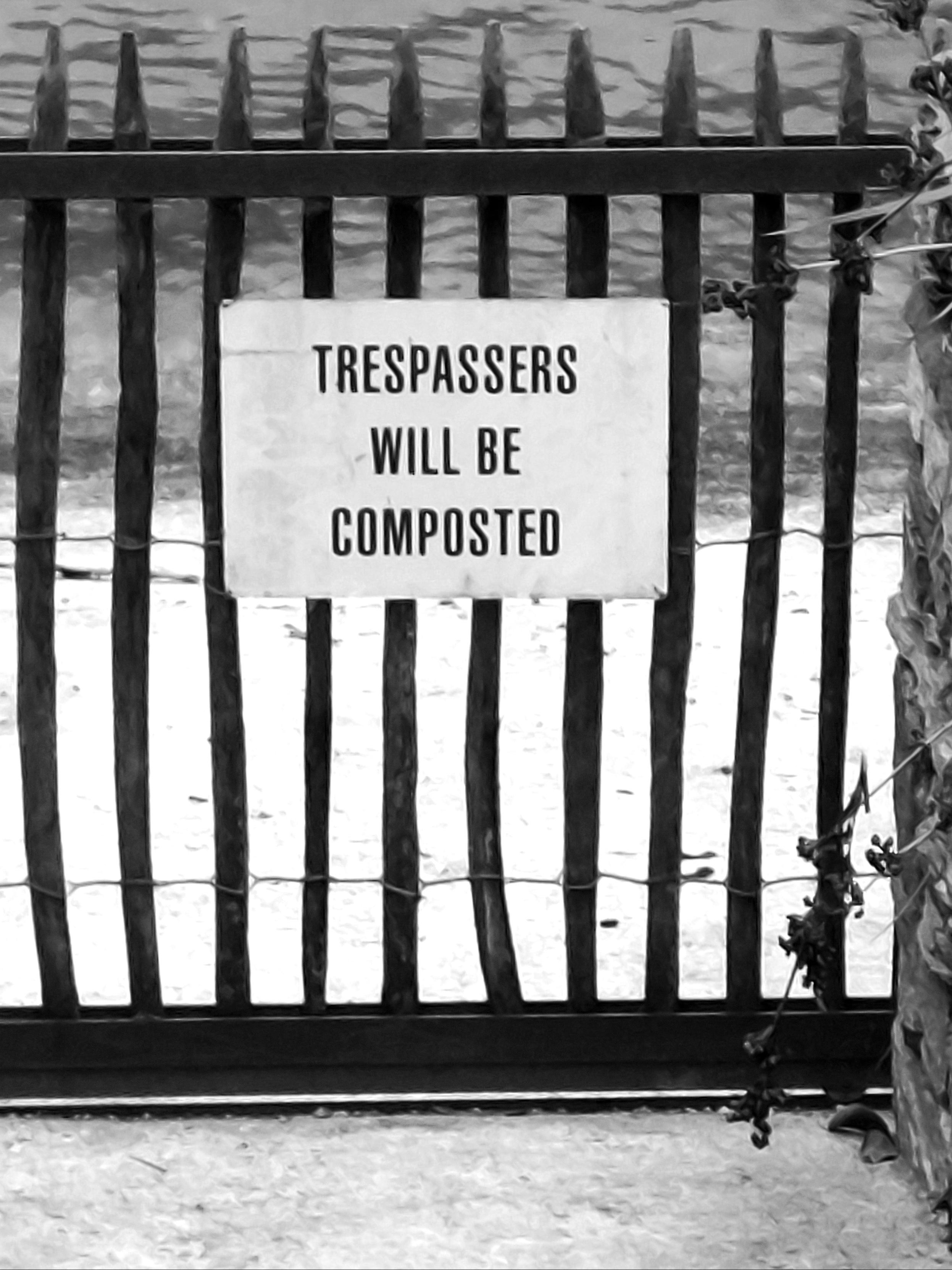 A white sign hangs on a picket fence, bearing the words ‘Trespassers will be composted’. In the background, sand and water.
 //  An einem Lattenzaun hängt ein weisses Schild mit der Aufschrift 'Trespassers will be composted'. Im Hintergrund Sand und Wasser.