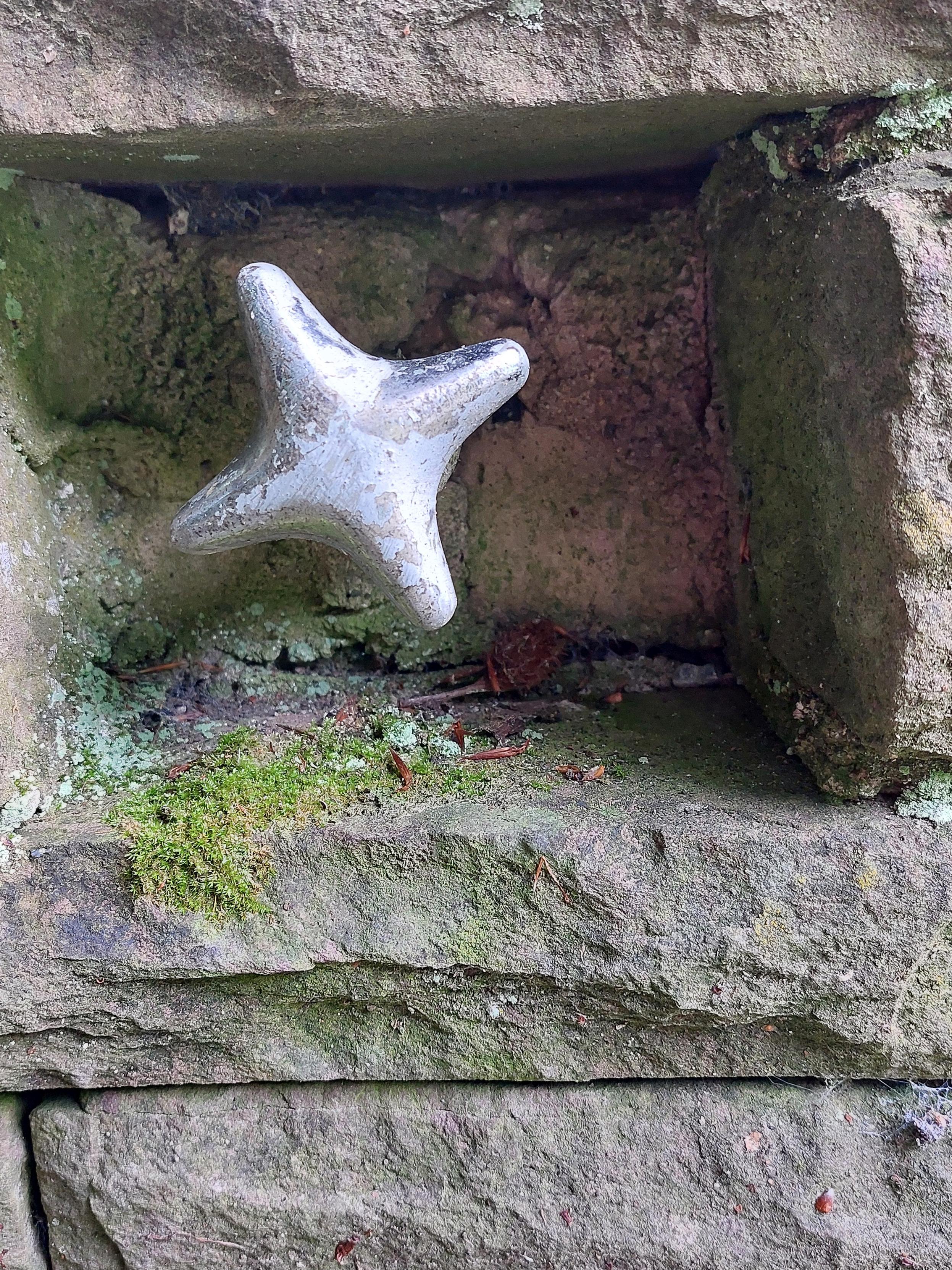 Close-up: A silver-coloured metal handle on a water valve. The slightly weathered handle is shaped like a cross with rounded corners. It is set into a rectangular recess made of reddish-grey sandstone, on which moss has already taken hold.